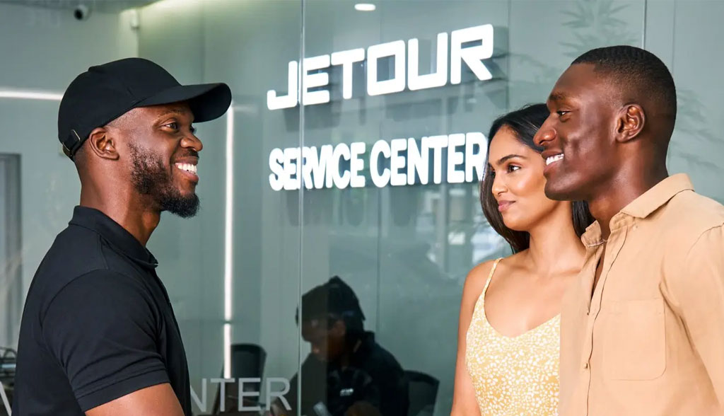 A man in a black cap greets a smiling couple at a service center. The atmosphere is friendly, with a "JETOUR SERVICE CENTER" sign in the background.