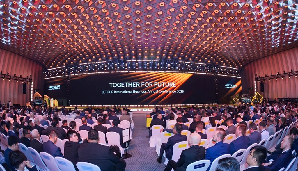 A large conference hall filled with attendees facing a stage displaying "TOGETHER FOR FUTURE" at the "JETOUR International Business Annual Conference 2025." The ceiling is adorned with geometric patterns, creating an elegant and professional atmosphere.