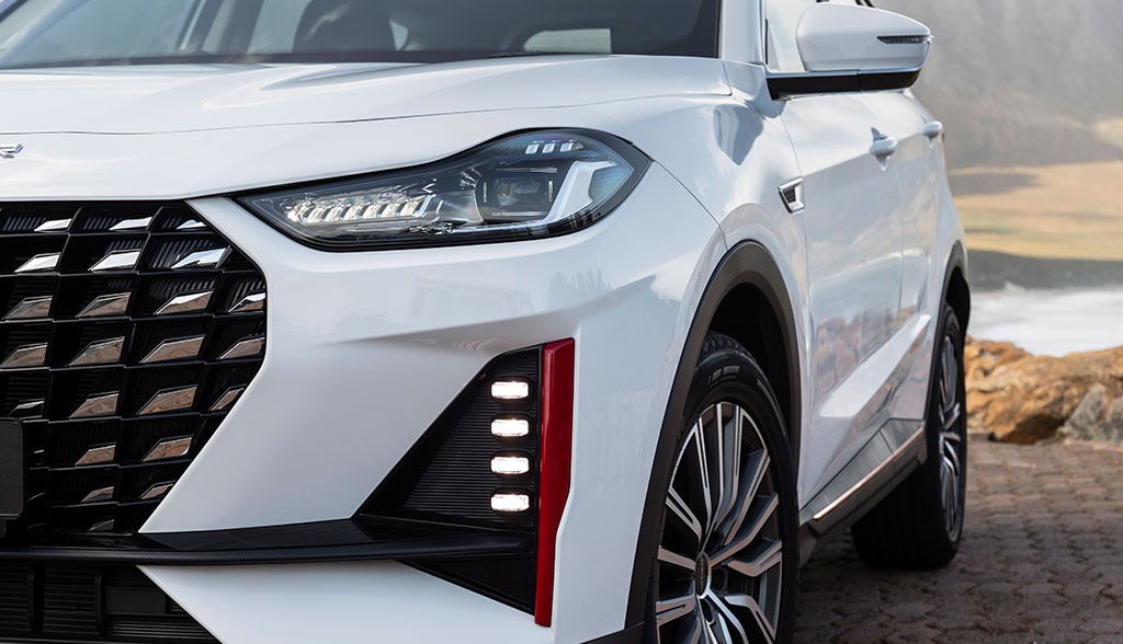 Close-up of a sleek, white SUV showcasing a modern headlight design, bold grille, and sharp body lines, parked on a cobblestone path by the sea.